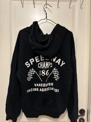Svart hoodie från h&m - Svart hoodie med dragkedja och huva. På ryggen står det 'Speedway Champs' och 'Vancouver Racing Association' med vita tryck och rutiga flaggor. Framtill finns en mindre Speedway Champs-logga. Köpt för många år sedan men knappt använd.