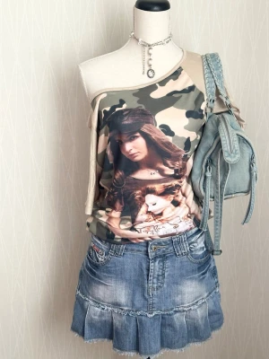 Camouflage topp med tryck - Trendig offshoulder topp med camouflage-mönster i gröna och beige toner. Framsidan har ett stort tryck med en tjej i keps och docka. Toppen är ärmlös på ena sidan och har en lös passform, perfekt för en cool och avslappnad stil.