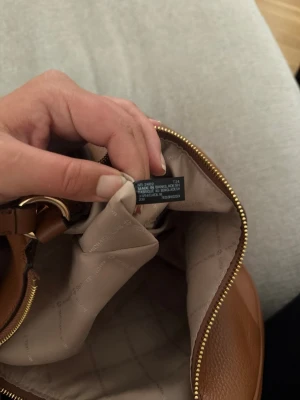 Brun handväska från Michael Kors - Snygg brun handväska från Michael Kors i äkta läder. Väskan har guldfärgade metalldetaljer, dragkedja och ett ljust, mönstrat innerfoder med flera fack. Perfekt för dig som vill ha en stilren och klassisk accessoar.