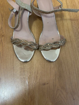 Glittriga guldiga pumps med blockklack - Säljer ett par glittriga pumps i guld med flätad rem framtill och justerbar ankelrem. Skorna har en hög blockklack dekorerad med stenar och metallicfinish. Perfekta för att sticka ut på festen! Materialet är syntet med glitterdetaljer.