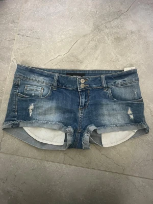 Jeansshorts - Säljer ett par blå denimshorts med råa kanter och slitna detaljer. Shortsen har fem fickor, bälteshällor och stängs med dragkedja och dubbla knappar. Perfekta för varma dagar och ger en avslappnad look.