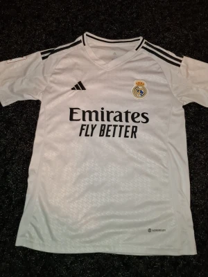 Real Madrid matchtröja vit Adidas - Säljer en vit Real Madrid matchtröja från Adidas med svarta detaljer och klubbmärke på bröstet. Tröjan har V-ringning, svarta ränder på axlarna och texten 'Emirates Fly Better' på framsidan. Tillverkad i lätt och ventilerande material.