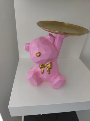 Rosa björn smyckesfat med guld - Snyggt smyckesfat i form av en geometrisk rosa björn med guldfärgad nos och rosett. Björnen håller upp ett löstagbart guldfärgat fat, perfekt för att förvara ringar, örhängen eller andra små accessoarer. Materialet är hårdplast och metall.