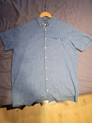 Blå linneskjorta från Dressmann, strl M - Snygg blå kortärmad skjorta från Dressmann i linnemix. Skjortan har klassisk krage, knappar framtill och en bröstficka med knapp. Perfekt för en avslappnad och stilren look.