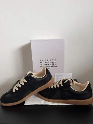 Maison Margiela svarta sneakers - Svarta sneakers från Maison Margiela med beige detaljer och snörning. Skorna har en klassisk siluett, beige innersula och yttersula i gummi. Materialmix av mocka och läder ger en lyxig känsla. Perfekta för dig som gillar stilrena och trendiga sneakers.