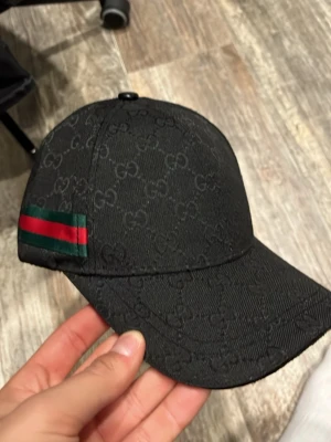 Svart keps med GG-mönster - Snygg svart keps med klassiskt GG-monogram över hela kepsen och en grön-röd rand på sidan. Justerbar rem baktill och böjd skärm. Perfekt för dig som vill ha en stilren accessoar med lyxig känsla.