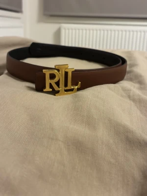 Brunt läderbälte med guldspänne - Snyggt brunt bälte i läder med ett stort guldfärgat spänne med bokstäverna 'RL'. Perfekt accessoar för att lyfta din outfit och ge en exklusiv touch. Bältet har klassisk design och justerbar längd.