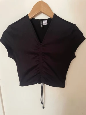 Svart croppad topp med snörning - Snygg svart croppad topp med v-ringning och snörning framtill. Kortärmad modell i ribbad stretchig kvalitet som sitter tight🖤