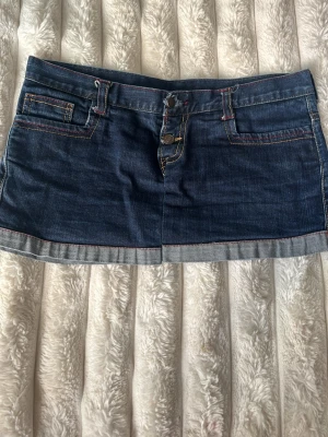 Mörkblå denimskort med kontrastsömmar - Snygg mörkblå denimskort med uppvikta kanter och kontrastsömmar i rött och gult. Klassisk femficksmodell med bälteshällor och knappgylf. Perfekt för en avslappnad och trendig look.