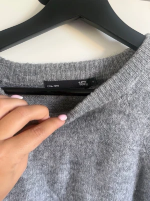 Grå stickad tröja från 157 - Mysig grå stickad tröja från 157 med rund halsringning och avslappnad passform. Tröjan har ribbstickade muddar vid hals, ärmslut och nederkant. Perfekt för lager på lager och enkel att matcha till olika outfits.