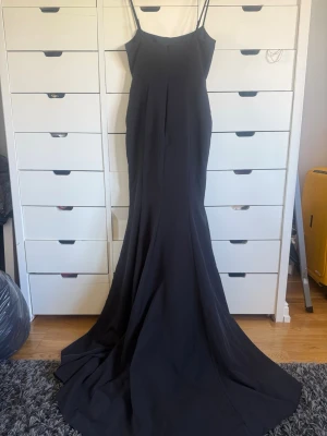 Svart långklänning - Elegant svart långklänning med smala axelband och figurnära passform. Klänningen har en lång, svepande kjol som ger en dramatisk look. Perfekt för speciella tillfällen där du vill sticka ut.