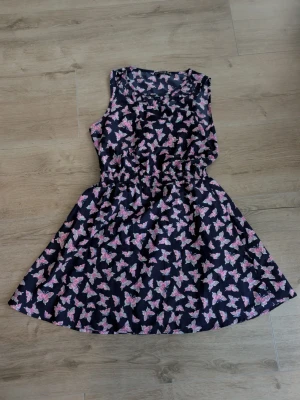 Mörkblå klänning med fjärilar - Supersöt mörkblå klänning från Primark med rosa och lila fjärilsmönster. Klänningen är ärmlös, har en markerad midja och en kort, utställd kjol. 