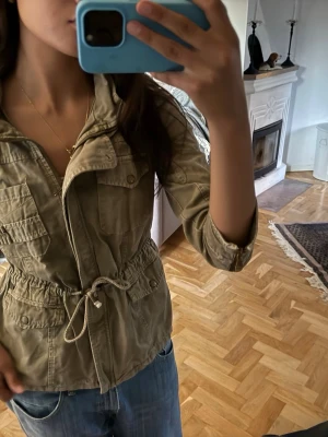 Beige parkajacka med snörning - Snygg beige parkajacka med dragsko i midjan och flera fickor framtill. Jackan har dragkedja och knappar, samt långa ärmar som kan vikas upp. Perfekt för vår och höst, och passar till jeans eller andra byxor.