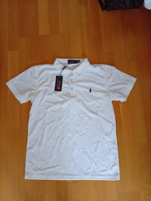 Vit pikétröja Ralph Lauren  - Klassisk vit pikétröja med krage och knappar framtill. Tröjan har korta ärmar och en svart broderad logga på bröstet. Perfekt till jeans eller shorts för en clean och stilren look.