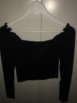 Svart offshoulder topp med långa ärmar - Trendig svart offshoulder topp med långa ärmar och ribbad struktur. Toppen har knyt på axlarna och en croppad passform som ger en snygg siluett. Perfekt att styla med höga jeans eller kjol för en cool look.