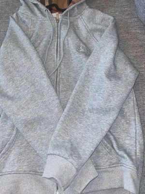 Ljusgrå zip hoodie med huva - Snygg ljusgrå hoodie med dragkedja och huva. Hoodien har dragsko i huvan, ribbade muddar och fickor framtill. Insidan av huvan har ett rutigt mönster i beige, brunt och rött. Materialet är mjukt och skönt, perfekt för chill dagar.
