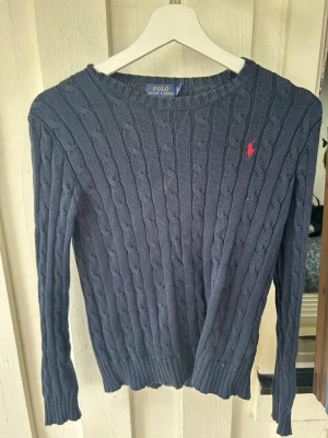 Marinblå kabelstickad tröja Polo - Kabelstickad marinblå tröja från Polo Ralph Lauren med rund halsringning och röd broderad logga på bröstet. Tröjan har långa ärmar och ribbade muddar vid ärmslut och nederkant. Perfekt för en klassisk och stilren look.
