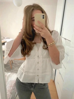 Vit blus med spetsdetaljer - Söt vit blus med knappar framtill och fina spetsdetaljer längs bröstet och ärmarna. Blusen har en lätt v-ringning och trekvartsärmar som ger en avslappnad men ändå chic look. Tillverkad i ett tunt, luftigt tyg som passar perfekt till jeans.