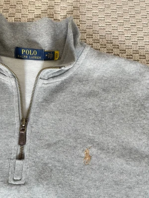 Grå halvzip från Polo Ralph Lauren - Snygg grå halvzip-tröja från Polo Ralph Lauren med klassisk broderad logga på bröstet. Tröjan har hög krage, dragkedja med läderdetalj och är tillverkad i mjukt bomullsmaterial. Perfekt för lager på lager och stilren look.