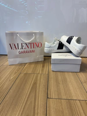 Valentino Garavani  - Snygga vita sneakers från Valentino Garavani med svart band över sidan och ikoniska nitar på sulan. Skorna har en klassisk låg profil och är tillverkade i slätt läder med vita snören. Perfekta för dig som vill ha en clean och exklusiv look.
