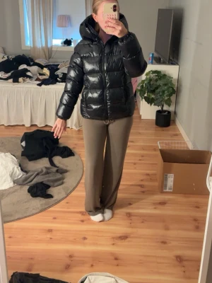 Svart glansig pufferjacka med huva - Säljer en svart, glansig pufferjacka med stor huva och dragkedja framtill. Jackan har en boxig passform och breda kanaler som ger en riktigt puffig look. Perfekt för kalla vinterdagar.