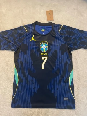 Blå Brasilien fotbollströja vinicius - Snygg blå fotbollströja för Brasilien med nummer 7 och VINI JR. på ryggen. Tröjan har gula och turkosa detaljer, CBF-emblem på bröstet och korta ärmar. Tillverkad i ett lätt och ventilerande material, perfekt för träning eller match.