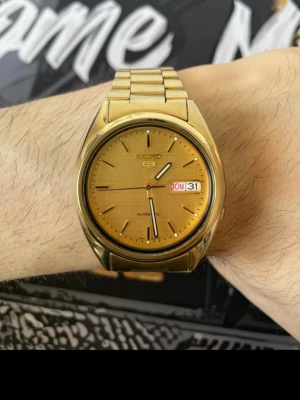 Guldfärgad Seiko 5 automatisk klocka - Stilren guldfärgad Seiko 5 automatisk klocka med metallarmband och dag- och datumvisning. Klockan har en rund urtavla i guldton med minimalistiska index och visare. Perfekt för dig som gillar klassisk och tidlös design.