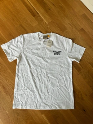 Gallery Dept. t-shirt - Säljer en vit t-shirt från Gallery Dept. med svart tryck fram och stort tryck på ryggen där det står 'GALLERY DEPT. HOLLYWOOD, CA.'. Klassisk passform och rund hals. Perfekt för dig som gillar streetwear och enkel stil.
