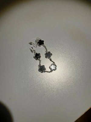 Silverarmband med svarta blommor - Snyggt armband i silverfärg med fem blommor i svart och en i pärlemor. Blommorna har en mjuk, rundad form och sitter på en tunn kedja med justerbar längd. Perfekt för dig som gillar stilrena och trendiga accessoarer.