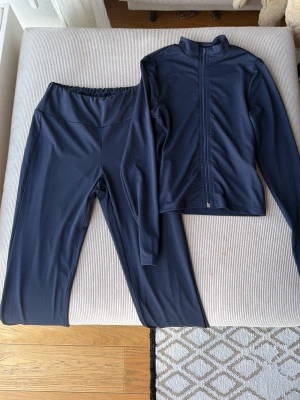 Mörkblått träningsset med dragkedja - Snyggt mörkblått träningsset med långärmad zip-jacka och matchande tights. Setet är gjort i ett stretchigt, mjukt material som passar perfekt för gym, löpning eller yoga. Jackan har hel dragkedja framtill och tightsen har bred midjeresår för extra komfort.