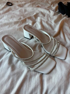 Klackskor/ balskor - Snygga vita pumps med fyrkantig tå och bred klack. Skorna har flera tunna remmar, varav några är täckta med glittriga detaljer som ger extra bling. Perfekta för dig som vill sticka ut och addera lite glamour till din outfit. Använda 1 gång på bal. Tror jag köpte dom från Nelly men är inte helt säker