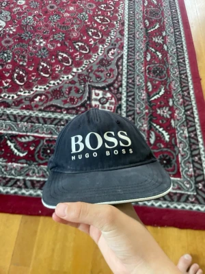 Svart keps från Hugo Boss - Snygg svart keps från Hugo Boss med klassisk böjd skärm och stor vit BOSS-logga framtill. Kepsen har ventilerande hål och justerbar passform bak. Tillverkad i bomullstyg som ger en avslappnad och stilren look.