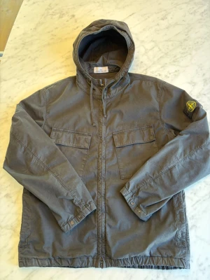 Svart short parka med luva från Stone Island - Snygg svart vindjacka från Stone Island med luva och dragkedja framtill. Modell Short parka. Jackan har två stora bröstfickor med lock och den klassiska Stone Island-loggan på vänster ärm. Perfekt för dig som gillar streetwear och vill ha en stilren look. Sparsamt använd. Nypris 6800 SEK. 