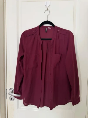 Vinröd blus från H&M Divided - Vinröd blus från H&M Divided med knappar framtill, två bröstfickor och axelklaffar. Blusen har en lös passform och är tillverkad i ett tunt, lätt material som ger ett snyggt fall. Perfekt att styla med jeans eller kjol.