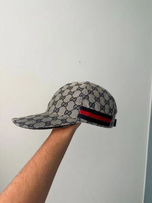 Gg keps navy grey - Säljer en GG keps, På sidan finns en röd och marinblå rand. Justerbar rem bak i svart läder. Perfekt för dig som vill ha en stilren och lyxig accessoar.