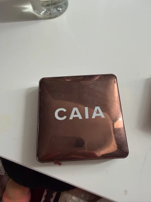 Bronzer från CAIA i metallisk dosa - Bronzer från CAIA i en fyrkantig, metallisk brun dosa med spegel i locket. Själva bronzern har en varm, skimrig brun ton och är pressad i rund form. Perfekt för att skapa glow och definition till ansiktet.