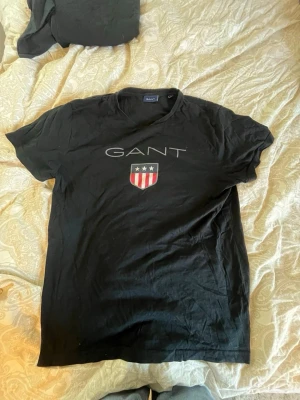Svart GANT t-shirt med tryck - Snygg svart t-shirt från GANT med stort tryck på bröstet och logga i vitt och rött. Klassisk passform och rund halsringning. Perfekt till jeans eller shorts för en avslappnad look.