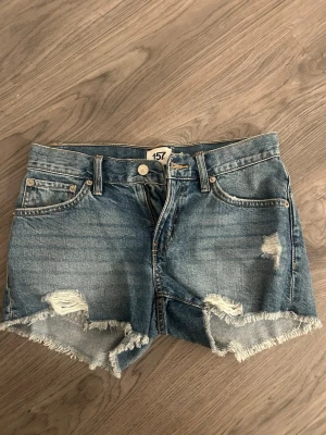 Blå jeansshorts med slitningar - Säljer ett par blå jeansshorts med råa kanter och slitna detaljer framtill. Shortsen har fem fickor, bälteshällor och knappgylf. Perfekta för en avslappnad och trendig look i sommar. Dessa är inte använda en enda gång 