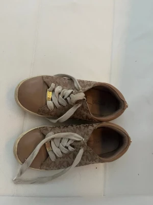 Bruna sneakers med logomönster - Snygga bruna sneakers från Michael Kors. Barn skor, 33-34 med beige logomönster på sidan och vita snören. Skorna har guldfärgade detaljer vid snörningen och en låg silhuett. Ovandelen är i textil och syntet, sulan är vit och beige.