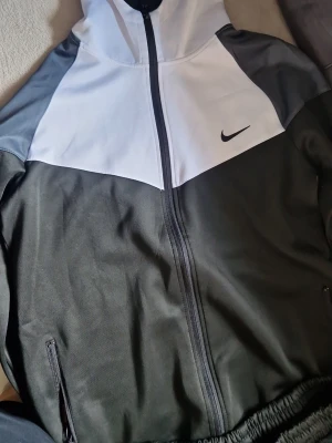 Nike svart och vit träningsdress - Snygg träningsdress från Nike med svart byxa och matchande jacka i svart, vitt och grått. Jackan har dragkedja, huva och två fickor. Byxorna är svarta med Nike-logga vid höften. Perfekt för träning eller chill.