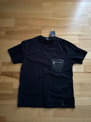 Svart Fendi t-shirt med ficka - Snygg svart t-shirt Fendi med korta ärmar och en unik bröstficka med dragkedja och Fendi-logga. Tillverkad i mjuk bomull och har en diskret Fendi-tag nedtill. Perfekt för dig som gillar exklusiva detaljer och stilren design.