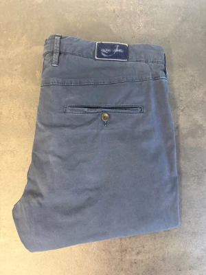 Blå chinos från Jacob Cohën, stl 35 - Snygga blå chinos från Jacob Cohën i klassisk rak modell. Byxorna har två sidofickor, bakficka med knapp och bälteshällor. Tillverkade i mjukt bomullstyg som ger en skön känsla och stilren look. Perfekta för dig som gillar en avslappnad men ändå trendig stil.