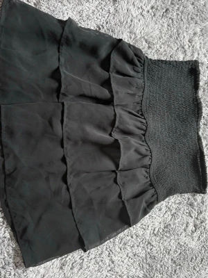 Svart volangkjol med smock - Säljer en svart kjol med bred smock i midjan och flera volanglager. Kjolen är gjord i ett tunt, mjukt tyg som ger ett flowy intryck. Perfekt för dig som gillar en feminin och trendig stil. Passar till många olika looks och är enkel att matcha.