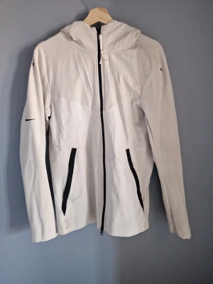 Beige/vit vindjacka/hoodie med huva från Nike - Vit vindjacka från Nike med hel dragkedja och två svarta fickor framtill. Jackan har huva, långa ärmar och diskreta svarta Nike-loggor på ärm och rygg. Materialet känns lätt och sportigt, perfekt för utomhusaktiviteter. Storlek S men passar bra på M också