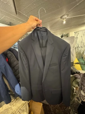 Mörkblå klassisk kavaj med byxor - Stilren mörkblå kavaj med två knappar framtill och klassiska slag. Kavajen har två fickor med lock och en bröstficka. Passar perfekt till skjorta och jeans för en snygg look. Materialet ser ut att vara en slät textil, troligen polyester eller ullblandning.