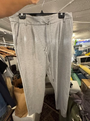Grå mjukisbyxor med dragsko - Snygga ljusgrå mjukisbyxor med dragsko i midjan och resår. Byxorna har fickor fram och dekorfickor bak. Tillverkade i mjukt och stretchigt material för maximal komfort. Perfekta för chill dagar eller när du vill ha en avslappnad stil. Finns att hämta i Gröndal.