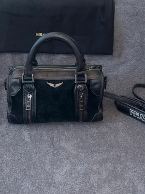 Zadig & Voltaire handväska - Cool svart handväska från Zadig & Voltaire i en mix av mocka och läder. Väskan har två rejäla handtag, avtagbar axelrem med logga och snygga silverdetaljer som dragkedjor och en ikonisk vinge framtill. Perfekt för dig som gillar rockig stil och vill ha något unikt.