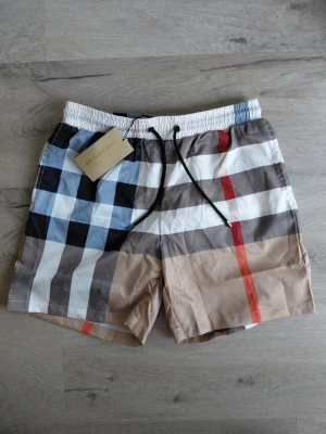  badshorts Burberry - Säljer ett par snygga badshorts från Burberry med klassiskt rutmönster i beige, vitt, svart, blått och rött. Shortsen har resår i midjan och svarta dragsnören med metalländar. Perfekta för stranden eller poolen.
