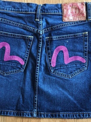 Kort jeanskjol med rosa detaljer - Denna jeanskjol från Evisu har en klassisk design med fem fickor och en knappgylf. Kjolen är i en mörkblå denimtvätt med kontrasterande gula sömmar. Baksidan pryds av de ikoniska måsvingelogotyperna i rosa på bakfickorna, samt en stor läderlapp med Evisu-logotypen i midjan. Litet märke på ena fickan (se foto).  Längd: 39 cm Bredd linning: 37 cm Vidd/nederkant: 48 cm
