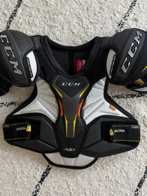 CCM Tacks AS1 axelskydd SR, storlek S - Säljer ett par svarta och vita axelskydd från CCM, modell Tacks AS1. Skydden har gula och orange detaljer samt vadderade axelpartier för extra skydd. Perfekta för hockey eller annan kontaktsport. Justerbara remmar för bästa passform.knappt använt. Anpassat för högsta ligan.  Nypris 2000-2500kr. 